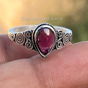 Könnte beinhalten: Ein Silberring mit einem zentralen, birnenförmigen, tiefroten Granat-Edelstein. Das Metallband ist auf beiden Seiten der Steinfassung mit komplizierten, geschwungenen Ornamenten verziert. Der Text SHIVANIGEMSCRAFTS ist im Hintergrund sichtbar.