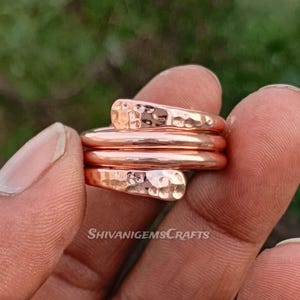 Puede incluir: Un conjunto de cuatro anillos de color cobre, uno envuelto alrededor de los otros. Los anillos superior e inferior tienen una textura martillada. Los anillos se sostienen en una mano. El texto "SHIVANIGEMSCRAFTS" es visible.