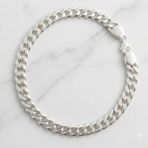Könnte beinhalten: Ein poliertes Panzerkettenarmband in Silberoptik mit ineinandergreifenden, flachen, ovalen Gliedern. Das Armband wird mit einem stabilen Karabinerverschluss gesichert und bildet eine durchgehende kreisförmige Schleife aus glänzenden Metallgliedern.