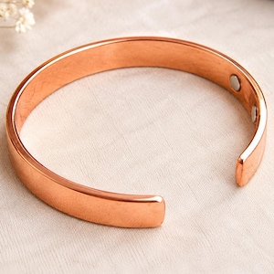 Könnte beinhalten: Ein poliertes, kupferfarbenes Armband in offener Form mit glatter Oberfläche. An der Innenseite des Armbands befinden sich zwei kleine, kreisförmige, silberfarbene Magnete, die nahe an den offenen Enden des Metallbandes angebracht sind.