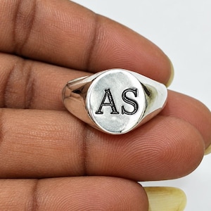 Könnte beinhalten: Ein polierter Siegelring in Silberoptik mit einer glatten, abgerundeten Oberfläche, in die die Buchstaben AS in einer klassischen Serifenschrift eingraviert sind. Der Ring hat ein massives, konisch zulaufendes Band mit gleichmäßigem Glanz.