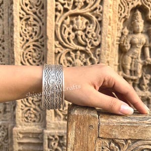 Könnte beinhalten: Ein silberfarbenes Armband aus Metall, bestehend aus mehreren schmalen, übereinanderliegenden Reifen mit komplexen, sich wiederholenden Prägemustern. Das Design zeigt parallele Linien sowie kleine florale oder geometrische Motive.