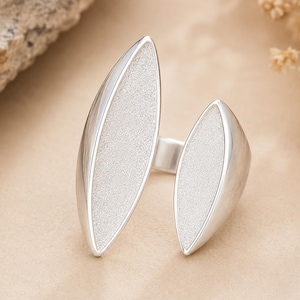 May include: A silver open ring featuring two leaf-shaped elements with a textured, matte finish on the front surfaces and smooth, polished edges. The band is open, connecting the two distinct, pointed shapes in a bypass style design.