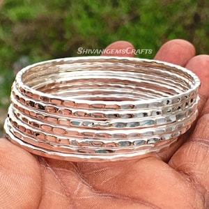 May include: A stack of silver bangles with a hammered texture. The bangles are round and appear to be made of metal. The image is taken outdoors with a blurred green background.