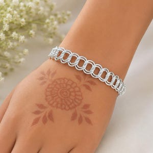 Könnte beinhalten: Ein silberfarbenes Armband mit einer Reihe von miteinander verbundenen, texturierten kreisförmigen Gliedern. Das Armband hat eine polierte, metallische Oberfläche und ein wiederkehrendes Muster aus Doppelringsegmenten.