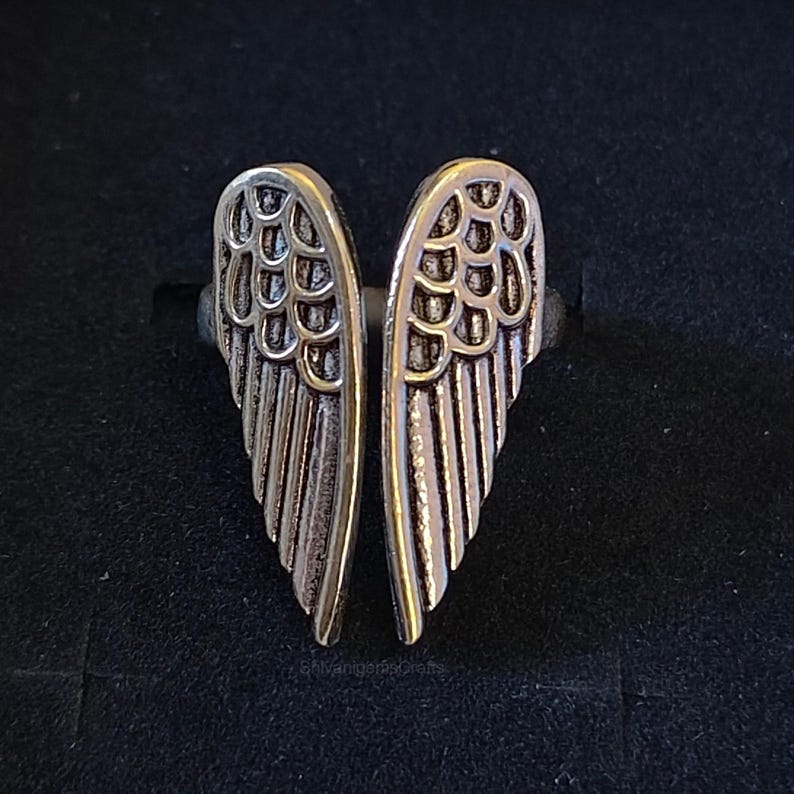May include: A silver-toned metal ring featuring two symmetrical angel wing designs that meet in the center. The wings have detailed, raised feather textures with darkened recessed areas to highlight the individual wing segments and structure.