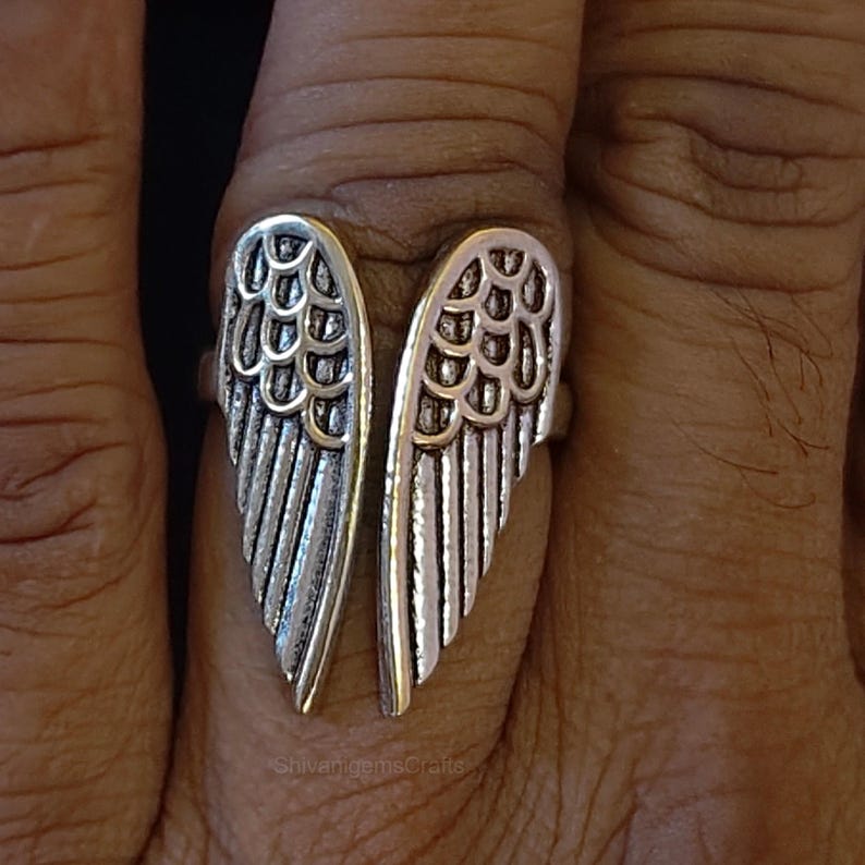 May include: A silver-toned metal ring featuring two symmetrical wing designs that meet in the center. The wings have detailed, layered feather textures with oxidized recessed areas to highlight the individual shapes. The text ShivaniGemsCrafts is visible.