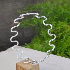 Könnte beinhalten: Ein silberfarbenes Armreif-Armband mit einem durchgehenden, wellenförmigen Design. Das Armband hat eine Reihe von Kurven und wird auf einem kleinen Holzblock präsentiert. Der Hintergrund ist grünes Laub. Der Text "ShivanigemsCrafts!" ist sichtbar.