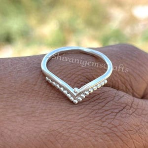 Könnte beinhalten: Ein silberfarbener Ring mit V-förmigem Design. Der Ring hat eine Reihe kleiner, runder Perlen entlang der Unterseite des V. Der Ring befindet sich auf einer braunen Oberfläche. Der Text "shivanigemcrafts" ist auf dem Ring sichtbar.