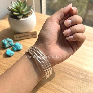 May include: A stack of thin, silver-colored bangles on a wrist. The image also shows turquoise stones, a small potted succulent, and the text "ShivaniGemsCrafts". The bangles are simple and elegant, suitable for everyday wear.