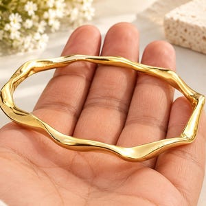 Könnte beinhalten: Ein polierter goldfarbener Armreif mit einem durchgehenden, unregelmäßig gewellten oder organisch gerippten Design. Die Metalloberfläche ist glatt und reflektierend und bildet eine fließende, ungleichmäßige Form als kreisförmige Schlaufe.