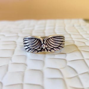 May include: A silver-toned metal ring featuring a detailed design of two symmetrical wings meeting at the center. The surface of the ring has an oxidized finish in the recessed areas to highlight the individual feather textures of the wings.