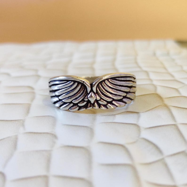 May include: A silver-toned metal ring featuring a detailed design of two symmetrical wings meeting at the center. The surface of the ring has an oxidized finish in the recessed areas to highlight the individual feather textures of the wings.