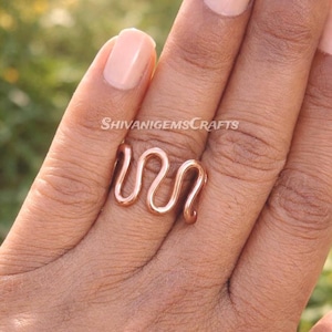 Könnte beinhalten: Ein kupferfarbener Ring mit einem wellenförmigen Design. Der Ring besteht aus einem einzigen Metallstück und hat eine glatte, polierte Oberfläche. Der Ring wird auf einem Finger gezeigt, mit dem Text "ShivanigemsCrafts" sichtbar.