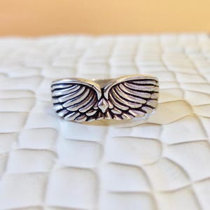May include: A silver-toned metal ring featuring a symmetrical design of two feathered wings meeting at a central diamond-shaped point. The band has a polished finish with darkened recessed areas to highlight the detailed texture of the feathers.