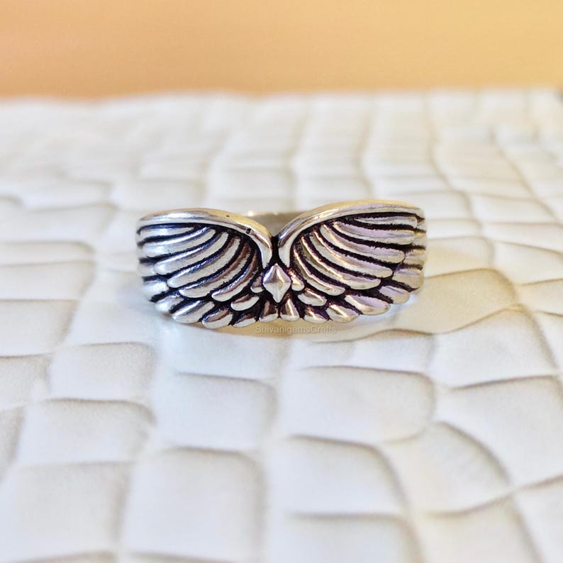 May include: A silver-toned metal ring featuring a symmetrical design of two feathered wings meeting at a central diamond-shaped point. The band has a polished finish with darkened recessed areas to highlight the detailed texture of the feathers.