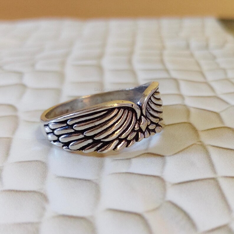 May include: A silver-toned metal ring featuring a detailed wing design that wraps around the band. The surface of the wings is textured with carved lines to represent individual feathers, with a small diamond-shaped accent at the center point.