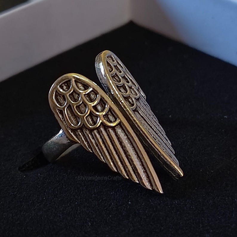 May include: A silver-toned metal ring featuring a detailed design of two symmetrical angel wings. The wings have textured, layered feather patterns with a darkened, oxidized finish in the recessed areas to highlight the intricate wing structure.