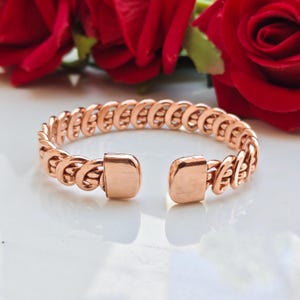 May include: A copper-colored metal cuff bracelet featuring a continuous woven or braided chain-link pattern. The bracelet has two smooth, flat, rectangular end caps that create an open gap, allowing the piece to be slipped onto the wrist.