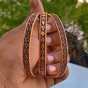 Könnte beinhalten: Drei kupferfarbene Metallarmreifen mit komplexen Prägemustern. Die äußeren Armreifen zeigen ein florales Motiv, während der mittlere Armreif ein geometrisches Design mit Rauten- und Blattformen aufweist. Der Text ShivanigemsCrafts ist sichtbar.