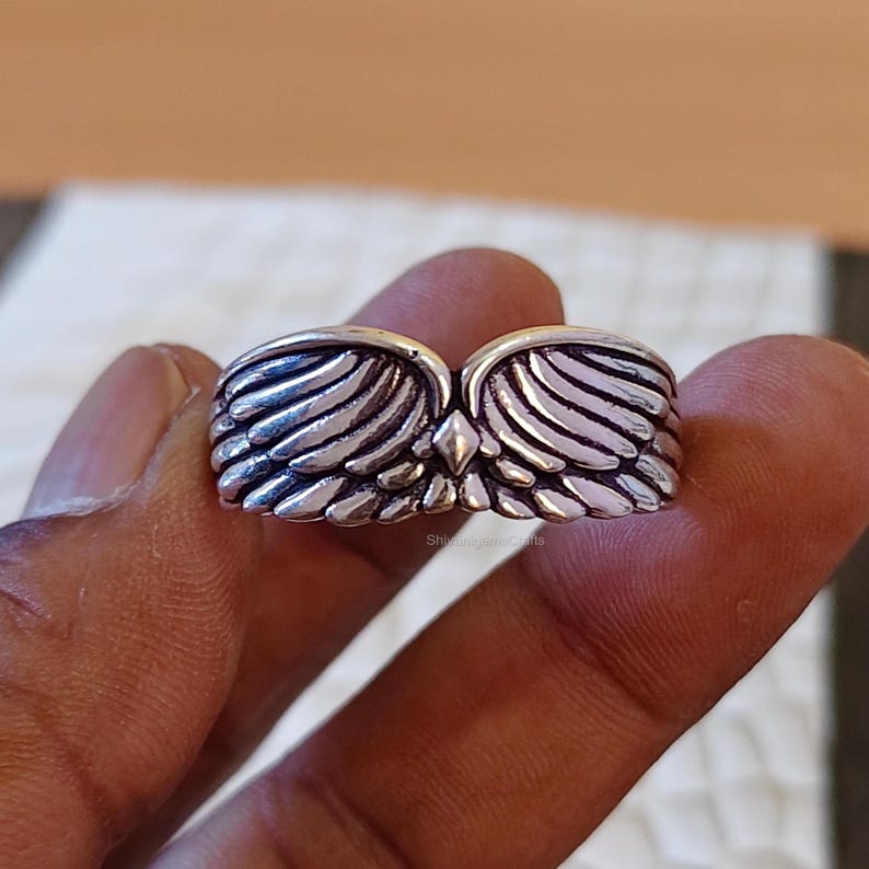 May include: A silver-toned ring featuring a symmetrical design of two feathered wings meeting at the center with a small diamond-shaped accent. The metal has a polished finish with darkened recessed areas to highlight the detailed wing texture.