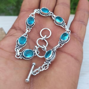 Könnte beinhalten: Ein silbernes Armband mit ovalen, hellblauen Edelsteinen. Das Armband hat einen Knebelverschluss und besteht aus miteinander verbundenen Gliedern. Jeder Edelstein ist in einem dekorativen, perlenbesetzten Rahmen gefasst. Das Armband wird auf einer Hand präsentiert.
