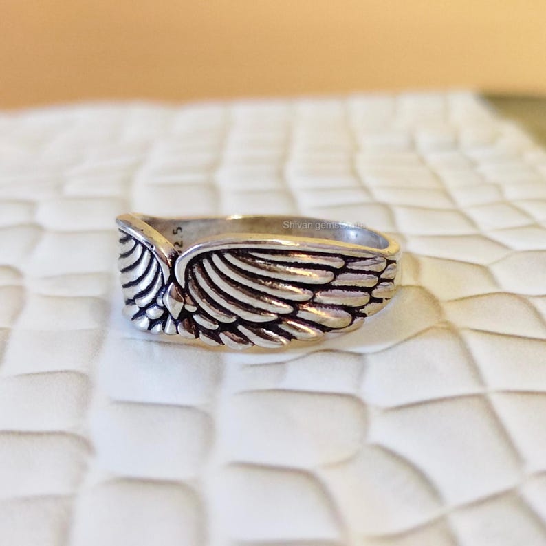 May include: A silver-toned metal ring featuring a detailed wing design that wraps around the band. The surface of the wings has deep grooves to define the individual feathers. The interior of the band shows the text 925 clearly visible.