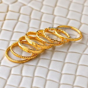 Könnte beinhalten: Ein Set aus fünf goldfarbenen Metallringen, die in einer diagonalen Reihe angeordnet sind. Jeder Ring weist eine eigene Textur auf, darunter glatte, gedrehte und facettierte Designs. Die Ringe sind dünn, kreisförmig und poliert.