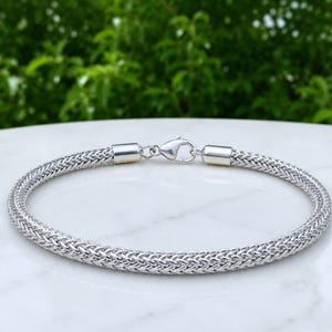 Könnte beinhalten: Ein silberfarbenes Weizenkettenarmband mit einem gewebten, geflochtenen Metallglieddesign. Das Armband wird mit einem polierten Karabinerverschluss und glatten zylindrischen Endkappen gesichert, was ein durchgehendes, flexibles Metallband bildet.