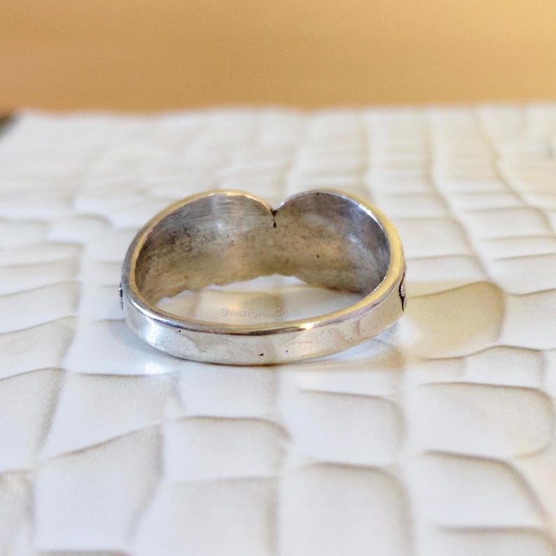 May include: A silver-toned metal ring featuring a smooth, polished band that curves upward into a central point. The interior of the band displays a subtle, etched texture, and the overall finish of the metal appears slightly weathered and matte.