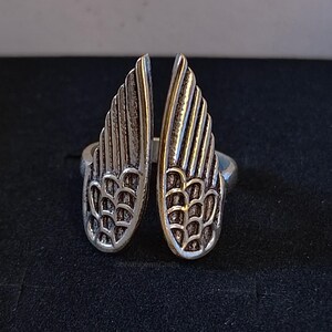 May include: A silver-toned metal ring featuring two symmetrical wing-shaped elements that meet at the center. The wings have detailed, etched feather patterns with darkened recessed areas to highlight the texture of the individual feathers.