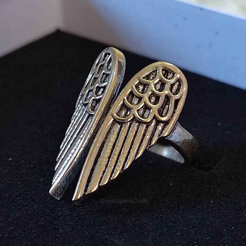 May include: A silver-toned metal ring featuring two symmetrical angel wing designs that meet at the center. The wings have detailed, raised feather textures with darkened recessed areas to highlight the intricate wing structure and shape.
