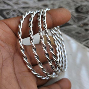 Könnte beinhalten: Eine Reihe silberfarbener Armreifen mit gedrehtem Design. Die Armreifen werden in einer Hand gehalten und zeigen ihre komplizierte, seilartige Textur. Das polierte Metall reflektiert das Licht und hebt die detaillierte Handwerkskunst des Schmucks hervor.