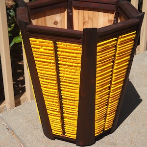 May include: A hexagonal wooden planter with a dark brown stain and yellow paint. The planter has a wide base and a narrow top.