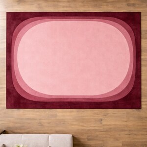 May include: A rectangular rug with a layered oval design in shades of pink and burgundy. The rug's central oval is a light pink, surrounded by progressively darker pink and burgundy borders. The rug is placed on a wooden floor.