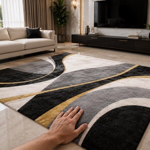 May include: A rectangular area rug with a modern design. The rug features a pattern of curved lines in black, gray, and cream, accented with gold. A hand is resting on the rug, showing its texture. The rug is in a living room setting.