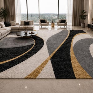 May include: A large area rug with a modern design. The rug features a pattern of black, gray, beige, and gold curved shapes. The rug is placed in a living room with a sectional sofa and large windows.