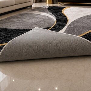 May include: A modern area rug with a geometric design. The rug features a combination of black, gray, and white colors with gold accents. The rug is partially rolled up, revealing the gray backing. The rug is placed on a light-colored tile floor.