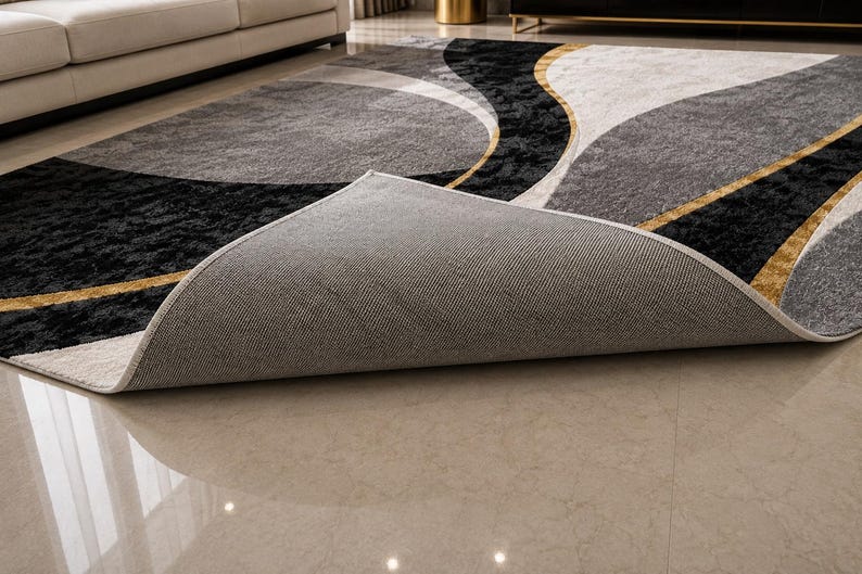 May include: A modern area rug with a geometric design. The rug features a combination of black, gray, and white colors with gold accents. The rug is partially rolled up, revealing the gray backing. The rug is placed on a light-colored tile floor.