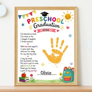 May include: A framed preschool graduation print with colourful text and illustrations. The print includes a handprint, the year 2026, and the name "Olivia". The design features a graduation cap, sun, hearts, stars, and a backpack.