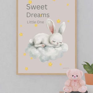 Puede incluir: Un estampado enmarcado con un conejo blanco durmiendo sobre una nube, con el texto "Sweet Dreams Little One". Estrellas amarillas adornan la imagen. Un osito de peluche rosa está sobre una mesita de noche.