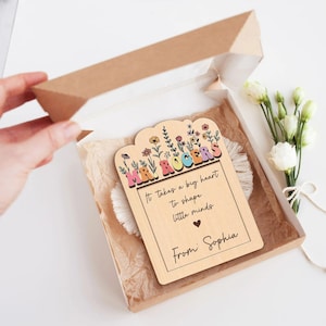 Puede incluir: Un regalo de agradecimiento para profesores de madera en una caja de papel kraft. El regalo presenta un diseño floral con el nombre "Mr. Rogers" y la cita "It takes a big heart to shape little minds". El regalo está personalizado con el nombre "Sophia".