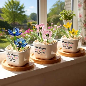 Puede incluir: Tres pequeños maceteros cuadrados de cerámica blanca con esculturas de flores de cristal. Cada macetero se asienta sobre una base redonda de madera. Las flores son azules, rosas y amarillas. El texto en los maceteros dice "gracias" y un mensaje personalizado.