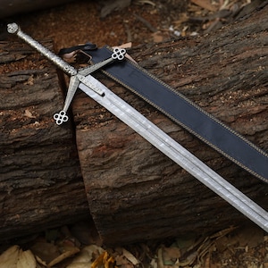Pode incluir: Uma espada longa e prateada, com punho e guarda ornamentados, repousa sobre um tronco desgastado. A espada tem uma lâmina texturizada e é acompanhada por uma bainha de couro preta. Sugere um tema histórico ou de fantasia.