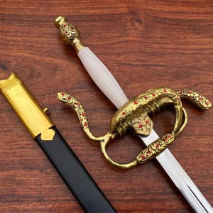 Pode incluir: Uma espada ornamentada com uma guarda dourada e um cabo branco. A guarda apresenta um design de cobra com detalhes vermelhos. A bainha é preta com detalhes dourados. A espada é exibida em uma superfície de madeira escura.