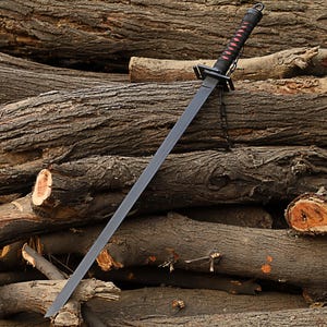 Puede incluir: Una espada katana negra con un mango rojo y negro, apoyada sobre una pila de troncos marrones. La hoja es recta y oscura, con una guarda rectangular. El mango está envuelto en material negro con detalles rojos.