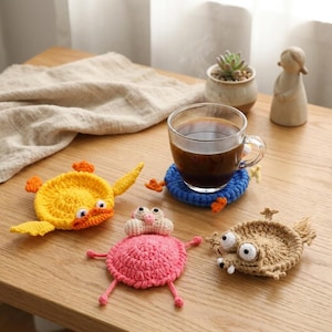 Könnte beinhalten: Eine Kaffeetasse steht auf einem blauen Häkeluntersetzer. Drei handgefertigte Häkeluntersetzer in Form einer gelben Ente, eines rosa Frosches und eines beigefarbenen Wesens liegen auf einem Holztisch. Die Untersetzer sind dekorativ.