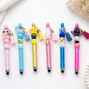 May include: Seven colourful stylus pens are displayed. Each pen has a unique design, such as a sloth, unicorn, star, ghost, and bat, with decorative beads and tassels. The pens are in shades of pink, blue, and yellow.