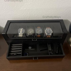 May include: A black rectangular watch storage box with a glass lid, containing four wristwatches. The box features a pull-out drawer with compartments holding a Sesto Synergy card, a small black box, watch links, and a blue ring.