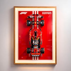 以下が含まれることがあります： 赤いフェラーリF1レーシングカーモデルの額装された画像。モデルは、白いストライプが入った赤い背景に配置されています。フェラーリのロゴとテキストが表示されています。フレームは木製です。